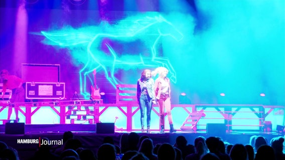 Auf einer Bühne tanzen zwei Sängerinnen verkleidet als Bibi und Tina vor einer bunten LED-Wand mit Neon-Symbolen. © Screenshot 