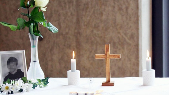 Auf einem Tisch mit weißer Tischdecke brennen neben einem kleinen Holzkreuz zwei kleinere weiße Kerzen, es stehen weiße Blumen in einer Vase auf dem Tisch sowie ein unkenntlich gemachtes. eingerahmtes Bild eines jungen Jugendlichen. © Screenshot 