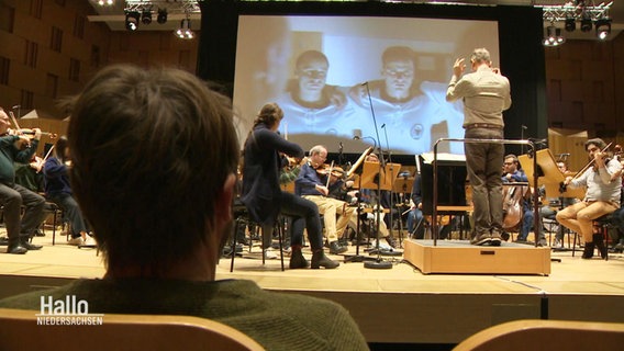In einem Konzertsaal sitzt ein Mann in der ersten Reihe und blickt auf ein probendes Orchester, das vor einer großen Leinwand mit Filmszenen spielt. © Screenshot 