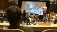 In einem Konzertsaal sitzt ein Mann in der ersten Reihe und blickt auf ein probendes Orchester, das vor einer großen Leinwand mit Filmszenen spielt. © Screenshot 