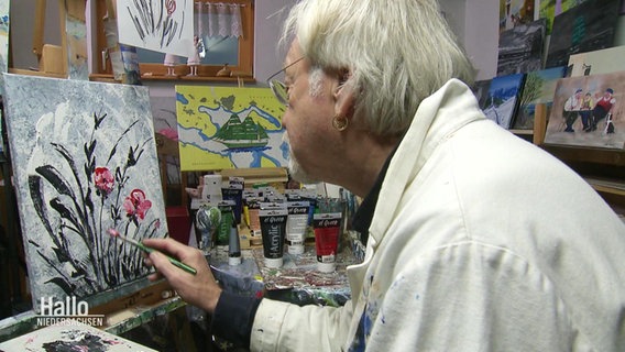 Ein älterer Herr sitzt konzentriert vorgebeugt in weißem Malerkittel vor einer Leinwand und malt ein Blumenbild in seinem privaten Atelier. © Screenshot 