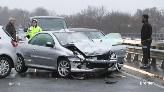 Auf einer Autobahnfahrbahn ist eine silberne Limousine nach einem Unfall komplett verbeult, ein Mann im Pullover steht daneben. © Screenshot 