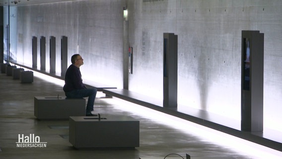 Ein Mann sitzt in einem langen Gang aus Betonwänden vor einem Bildschirm in einer Holocaust-Gedenkstätte. © Screenshot 