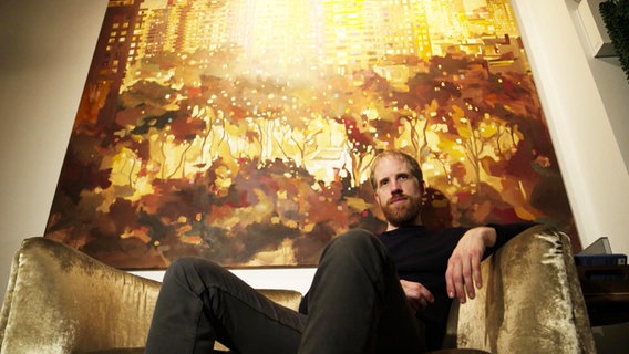 Ein junger Mann sitzt auf einem goldenen Sofa unter einem großem Gemälde. © Screenshot 