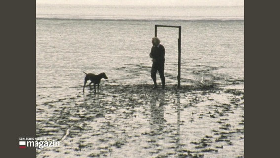 Bildhauer Hein Hoop, in Begleitung mit Hund, neben einer seiner Skulpturen im Watt. © Screenshot 