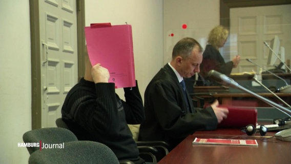 Ein Angeklagter hält sich im Gerichtssaal eine rote Mappe vor das Gesicht, neben ihm sitzt sein Verteidiger. © Screenshot 
