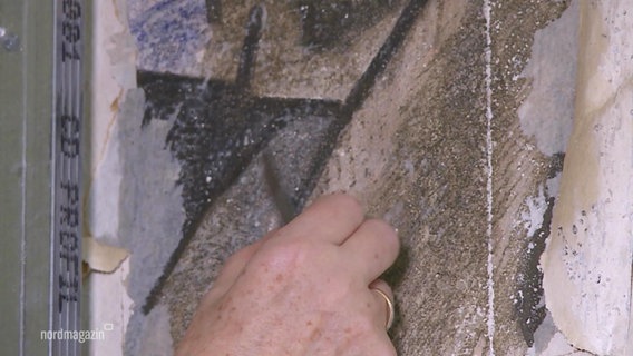 Eine Hand legt eine unter Weißer Farbe verstecktes Wandgemälde mit einem Kratzer frei. © Screenshot 