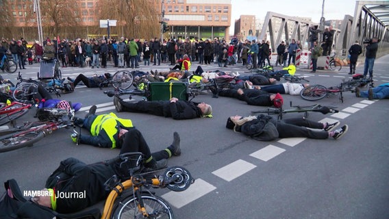 Viele Menschen liegen bei einer Mahnwache auf dem Rücken auf einer Straßenkreuzung. Neben ihnen liegen Fahrräder. Im Hintergrund stehen weitere Personen. © Screenshot 