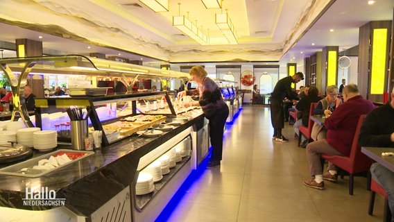 In einem asiatischen Restaurant steht eine Frau an der langen Buffet-Zeile in der Mitte des Essbereichs. © Screenshot 