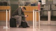 Ein Mann sitzt vor einem Flughafenschalter auf seinem Koffer und schaut auf sein Handy. © Screenshot 