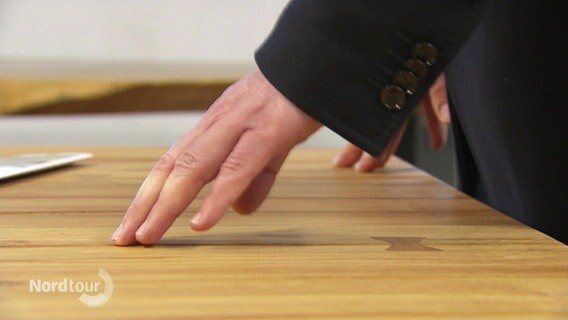 Eine Hand streicht auf der gemaserten Oberfläche eines Holztisches entlang. © Screenshot 