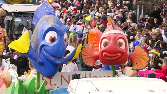 Zwei größere Fischfiguren im Zeichentrickstil sind auf einem Wagen bei einem Karnevalsumzug montiert. Im Hintergrund: zahlreiche Menschen stehen am Straßenrand. © Screenshot 