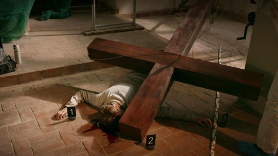 Ein Mann liegt mit dem Gesicht auf dem Boden einer Kirche. Unter seinem Kopf hat sich Blut gesammelt, auf seinem Körper liegt ein großes, umgekipptes Holzkreuz. © Screenshot 