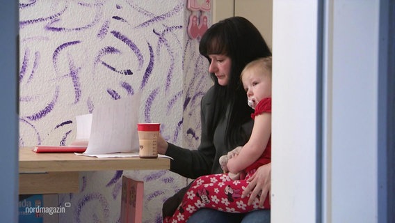 Eine Frau mit einem Kleinkind auf dem Schoß sitzt am Tisch und sieht Unterlagen durch. © Screenshot 