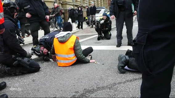 Ein Aktivist der Gruppe "Letzten Generation", der sich auf der Straße festgeklebt hat. Polizisten versuchen, seine Hände zu befreien. © Screenshot 