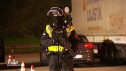Ein Polizist mit Maschinengewehr steht auf einem Autobahn-Parkplatz. © Screenshot 