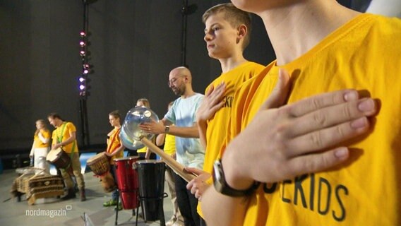 Mehrere Jugendliche stehen nebeneinander und haben die Hand auf die Brust gelegt. Einige spielen Trommel. © Screenshot 