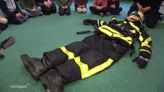 Eine Puppe in Feuerwehrausrüstung umzingelt von Kindern. © Screenshot 