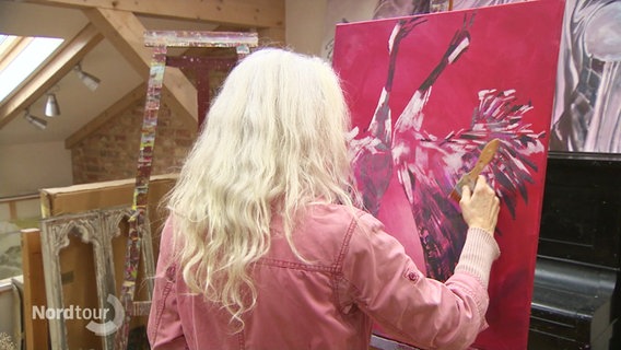 Eine Malerin malt ein rotes Bild mit Kranichen. Die Malerin ist von hinten zu sehen. © Screenshot 