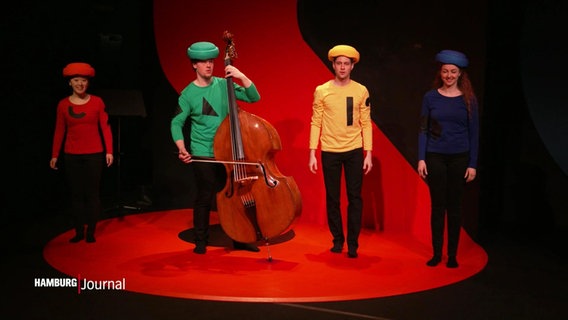 Vier Schauspielende auf einer Bühne. Sie sind entweder rot oder grün oder gelb oder blau gekleidet, einer spielt Kontrabass. © Screenshot 