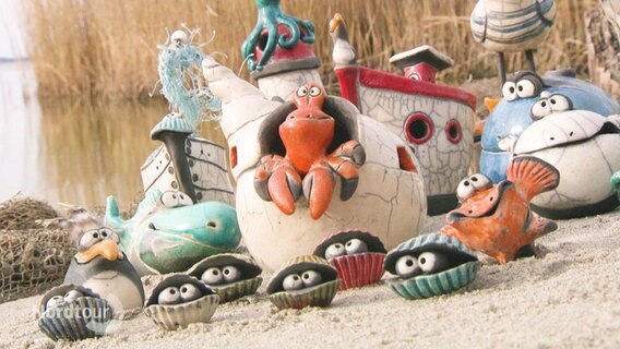 Mehrere bunt bemalte Keramik-Figuren mit maritimen Zügen sind auf einer Sandfläche an einem Ufer angeordnet. © Screenshot 