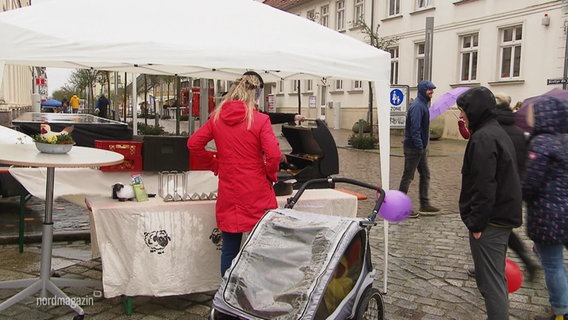 Auf einem verregneten Frühlingsfest stehen einige Menschen unter weißen Pavillion-Zelten. © Screenshot 