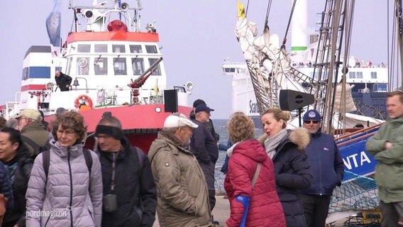 Mehrere Menschen stehen an einer Hafenkante, im Hintergrund sind Schiffe zu sehen. © Screenshot 