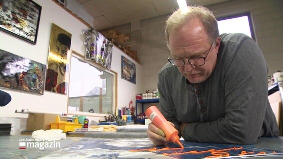 Ein Mann malt mit einer Spritzdose Farbe auf ein größeres Bild in einem Atelier. © Screenshot 