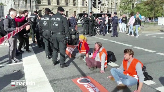 Mehrere junge Menschen sitzen auf einer Straße und blockieren den Verkehr. Vor ihnen steht Polizei. © Screenshot 