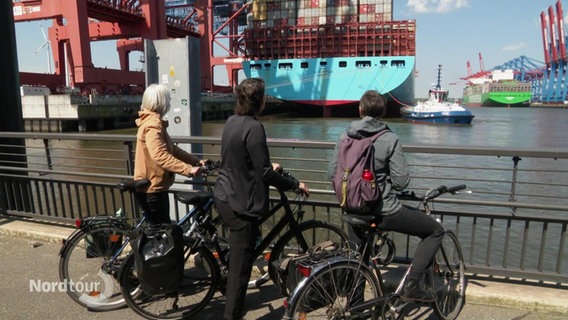 Drei Fahrradfahrerinnen gucken von der Hafenkante aus auf ein riesiges Container-Schiff, dass an einem Hamburger Container-Terminal vor Anker liegt. © Screenshot 