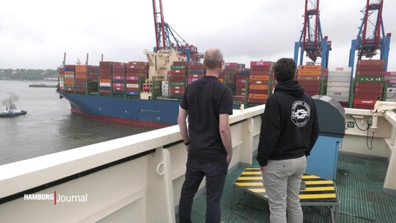 Zwei Männer stecken auf dem Deck eines Containerschiffes und schauen auf den Hamburger Hafen. © Screenshot 
