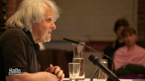 Peter Urban spricht in ein Mikrofon und liest aus seiner Biografie vor. Im Hintergrund ist verschwommen Publikum zu erkennen. © Screenshot 