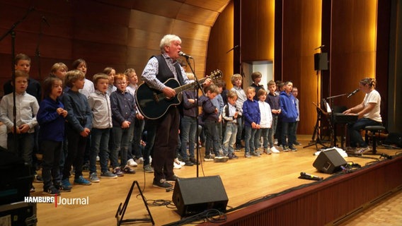 Rolf Zukowski steht mit Gitarre auf der Bühne und singt in ein Mikrophon, hinter ihm steht ein Chor mit Kindern. © Screenshot 