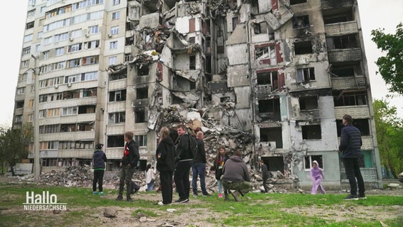 Eine  Gruppe an Menschen steht vor den Ruinen eines zerbombten Hochhauses in der Ukraine. © Screenshot 