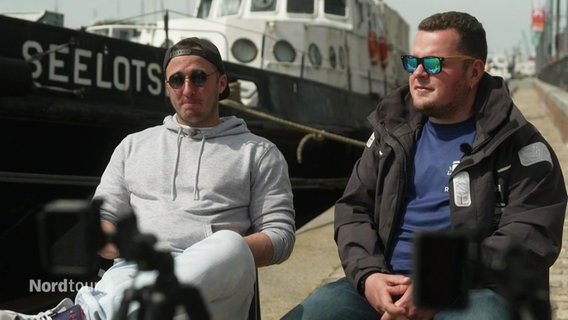 Zwei Männer mit Sonnenbrillen sitzen auf Stühlen an einem Hafenkai vor einer Kamera. © Screenshot 