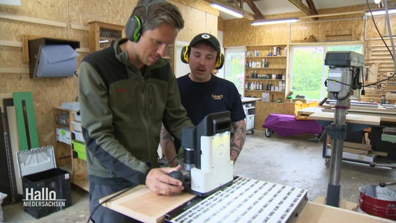 Moderator Arne-Torben Voigts bedient eine Holzfräse in einer Werkstadt, neben ihm steht ein Tischler begutachtend. © Screenshot 