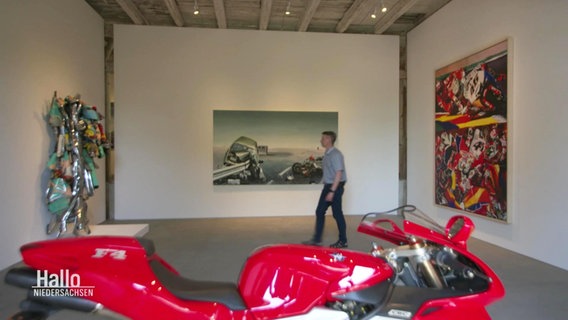 In einem Ausstellungsraum sind Bilder und ein Motorrad zu sehen. © Screenshot 