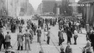 Historische Foto: Bei dem DDR-Volksaufstand vom 17. Juni 1953 stehen mehrere Menschen auf einem öffentlichen Platz, im Hintergrund fährt ein Panzer. © Screenshot 