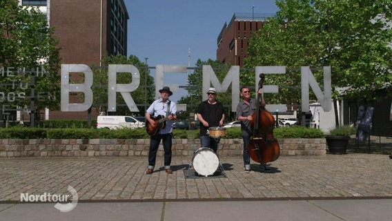 Vor vier großen Buchstaben, die zusammen das Wort "Bremen" ergeben, stehen drei Musiker auf einem öffentlichen Platz. © Screenshot 