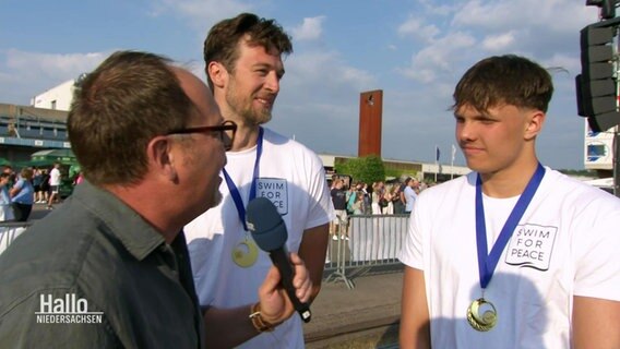 Ein NDR Reporter im Interview mit zwei Schwimmern mit Medaillen um den Hals. © Screenshot 