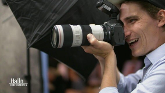 Ein Fotograf hält eine professionelle Spiegel-Reflex-Kamera mit langem Objektiv vor sein Auge. © Screenshot 