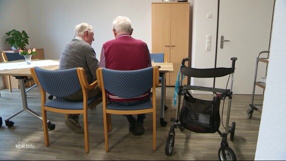 Zwei ältere Männer mit weißem Haar sitzen an einem Tisch, sie sind von hinten zu sehen. Neben ihnen steht ein Rollator. © Screenshot 