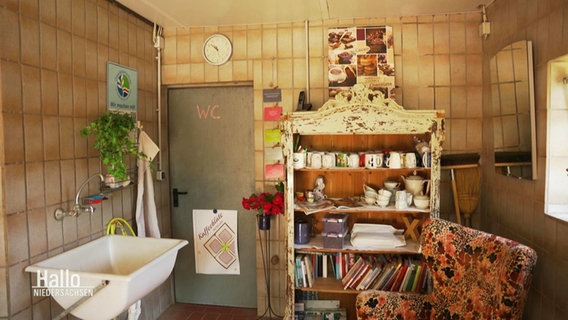 Blick in einen Raum mit befliesten Wänden: Neben einer Wassertränke stehen ein Sessel und ein liebevoll eingerichtetes Regal. © Screenshot 
