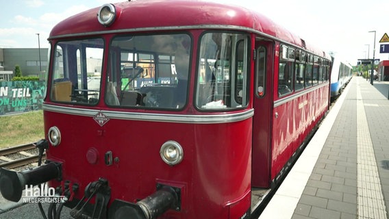 Eine historischer roter Zug-Waggon steht an der Spitze eines Regionalzugs an einem Bahnhof. © Screenshot 
