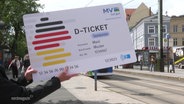 Ein großes Schild, das wie das Deutschlandticket für Senioren bedruckt ist, wird in die Kamera gehalte. Im Hintergrund eine Straßenbahnhaltestelle. © Screenshot 