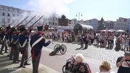 Auf einem öffentlichen Platz in Wismar geben als altertümlich Soldaten verkleidete Männer Salutschüsse ab. © Screenshot 