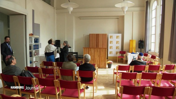 Blick in den Gebetsraum der jüdischen Gemeinde in St. Pauli in Hamburg. Rot gepolsterte Stühle stehen in Reihen hintereinander. Vereinzelt sitzen schon Menschen auf ihnen. © Screenshot 