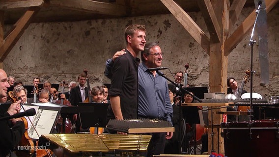 Zwei Männer stehen auf einer Bühne vor einem Orchester. © Screenshot 