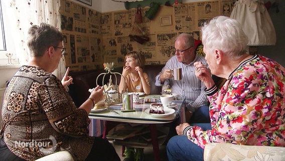 Menschen sitzen um einen Tisch und essen Kuchen in einem bunt dekoriertem Cafe. © Screenshot 