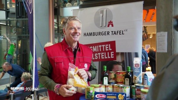 Moderator Jörg Pilawa posiert für ein Bild. Er trägt eine rote Weste der Hamburger Tafeln und hält eine Verpackung mit Aufbackbrötchen hoch. © Screenshot 